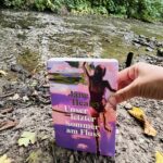 Buch Unser letzter Sommer am Fluss wird von einer Hand festgehalten. Im Hintergrund ist ein Fluss zu sehen.